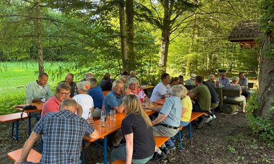 Menschen sitzen eng zusammen an Holzbänken, plaudern und trinken, umgeben von einem Wald mit grünen Bäumen und Wiese im Hintergrund. Es gibt keinen Text im Bild.