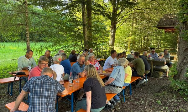 Menschen sitzen eng zusammen an Holzbänken, plaudern und trinken, umgeben von einem Wald mit grünen Bäumen und Wiese im Hintergrund. Es gibt keinen Text im Bild.